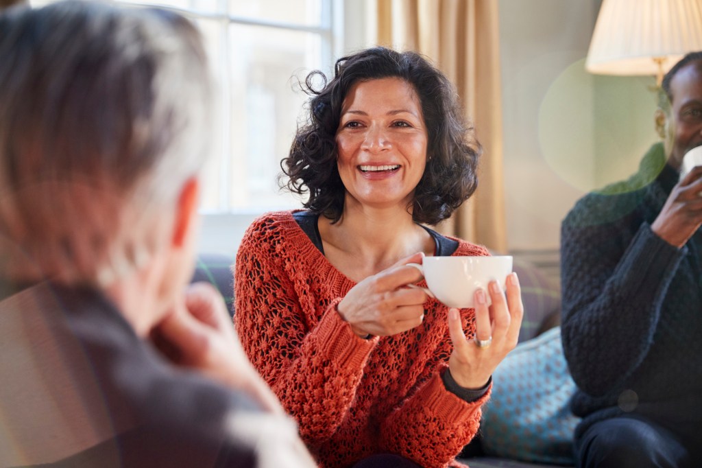 A middle-aged female sits on the couch with friends while holding a cup of tea.