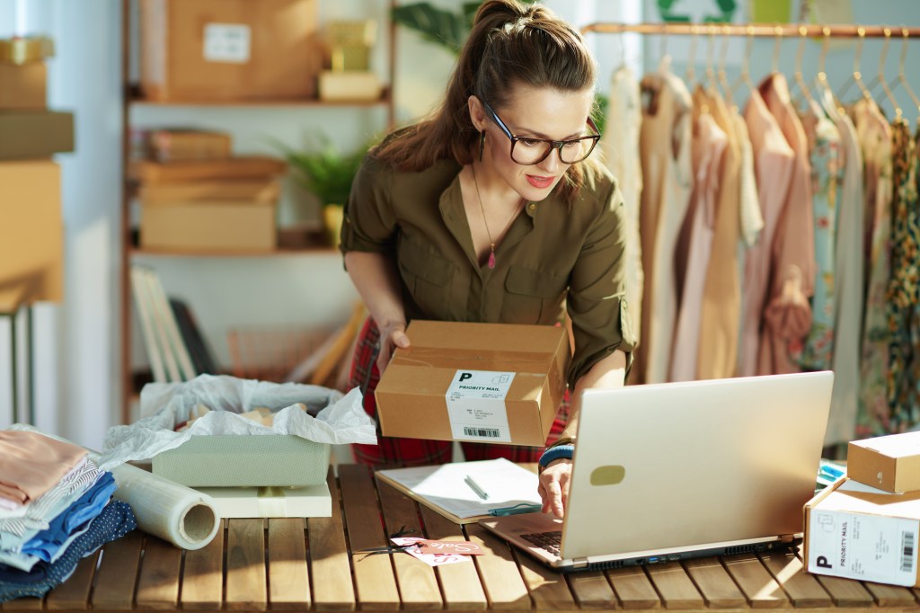 Middle aged small business owner woman with parcel using laptop in the office.