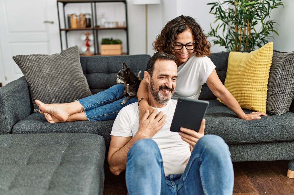 A man and woman smiling while looking down at a shared tablet. The house dog lounges on the female's lap.