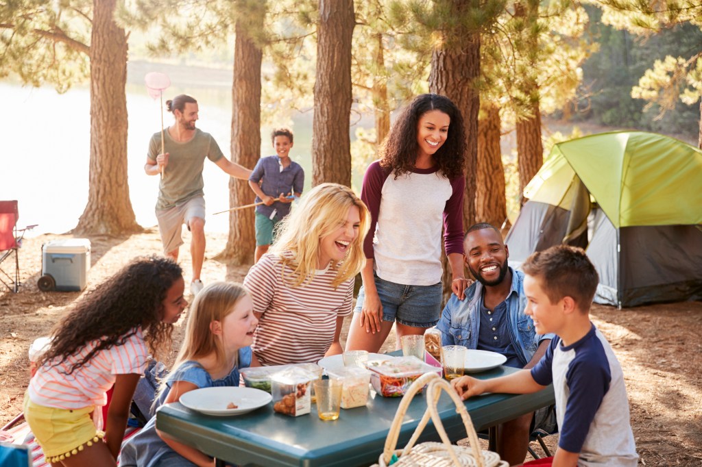 A group of friends and family laugh and stand around a picnic table at an outdoor campsite.