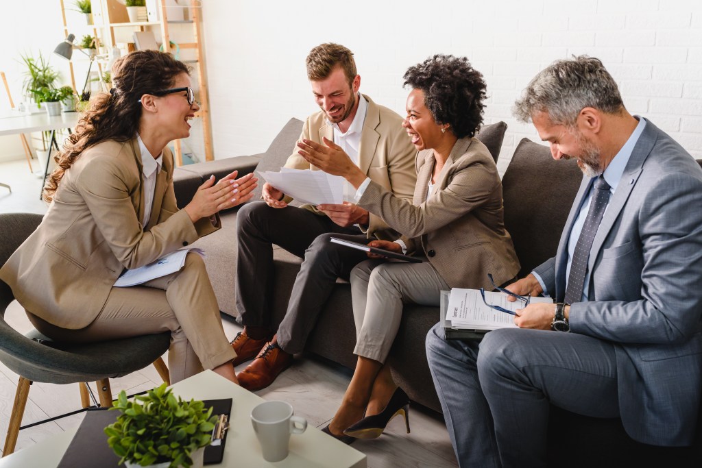 A group of four businesspeople sitting on a couch, laughing while looking at spreadsheets.