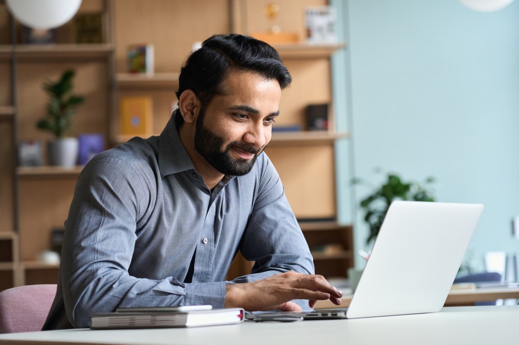 A middle-aged male sits at a table looking at his laptop and calendar.