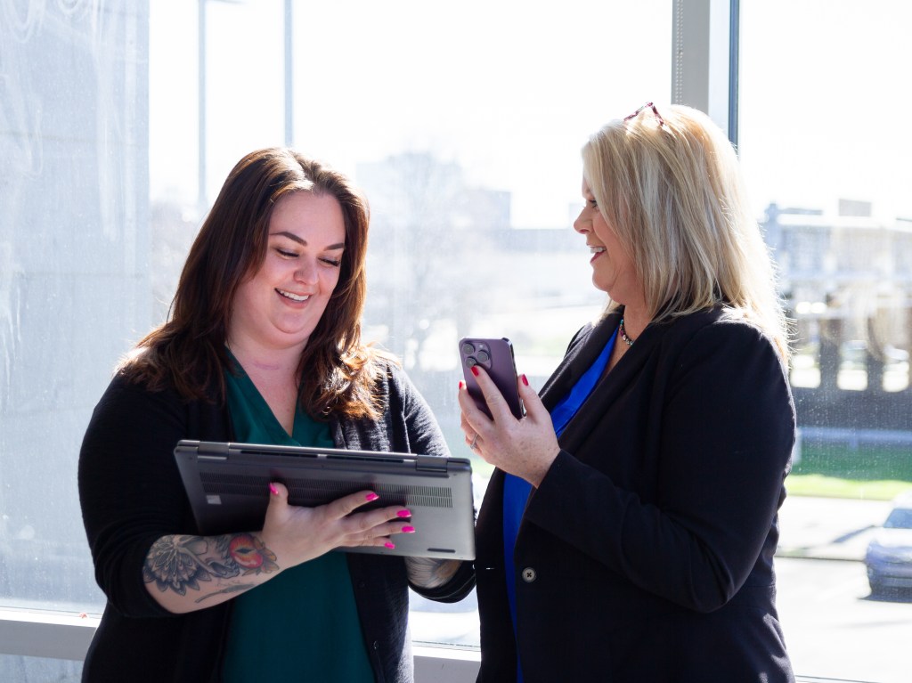 Two Peoples Bank employees stand smiling, one is on a portable laptop, and the other on her mobile phone.