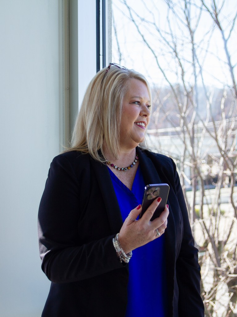 A woman stands holding her mobile phone while looking out a bay window.
