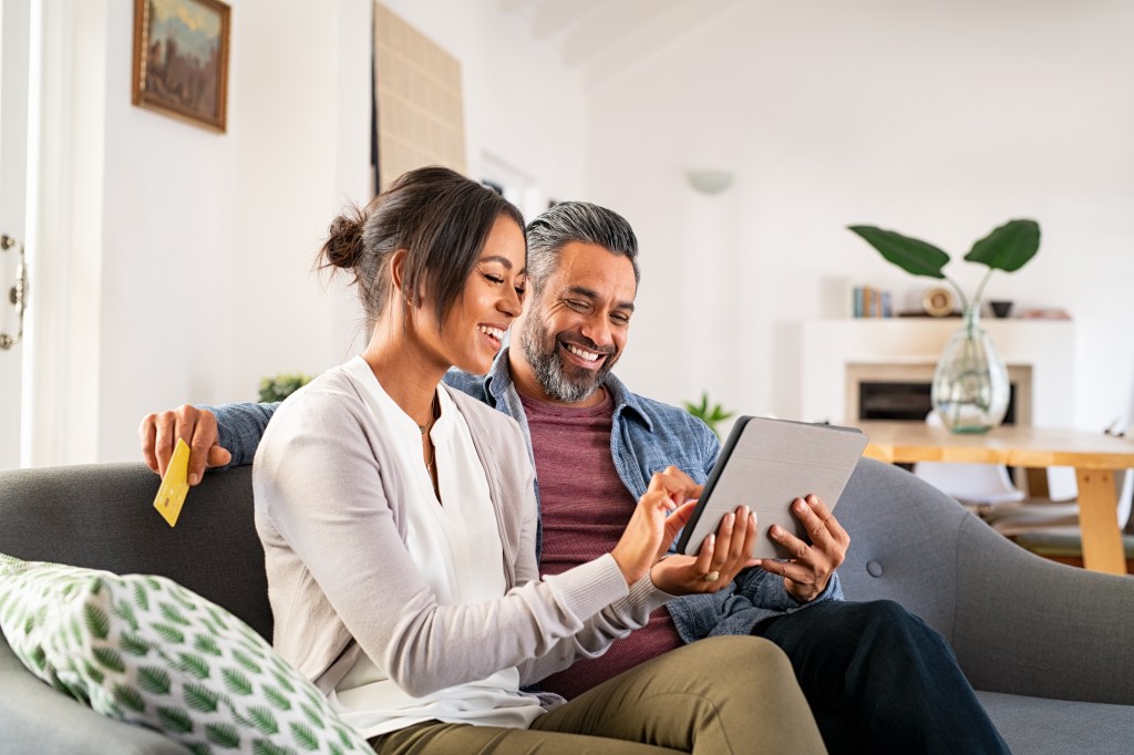 A couple sits on their couch smiling while looking at an iPad together.