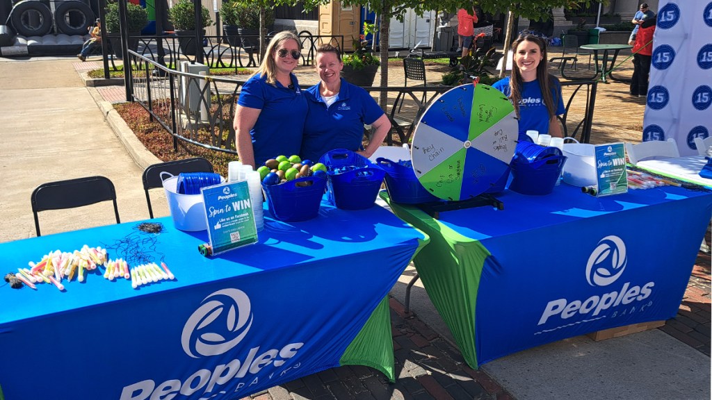 Peoples Bank employees stand at a booth they are hosting at the First Friday Fan Fest.