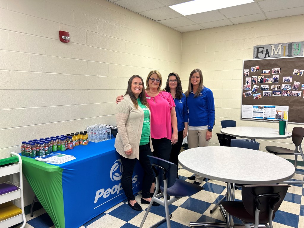 Four Peoples Bank employees stand in front of a table they prepared with drinks and snacks for local teachers.