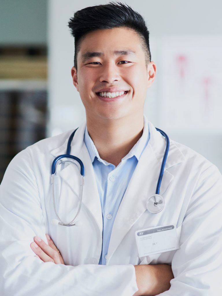 A young male doctor smiles directly at the camera with his arms crossed.