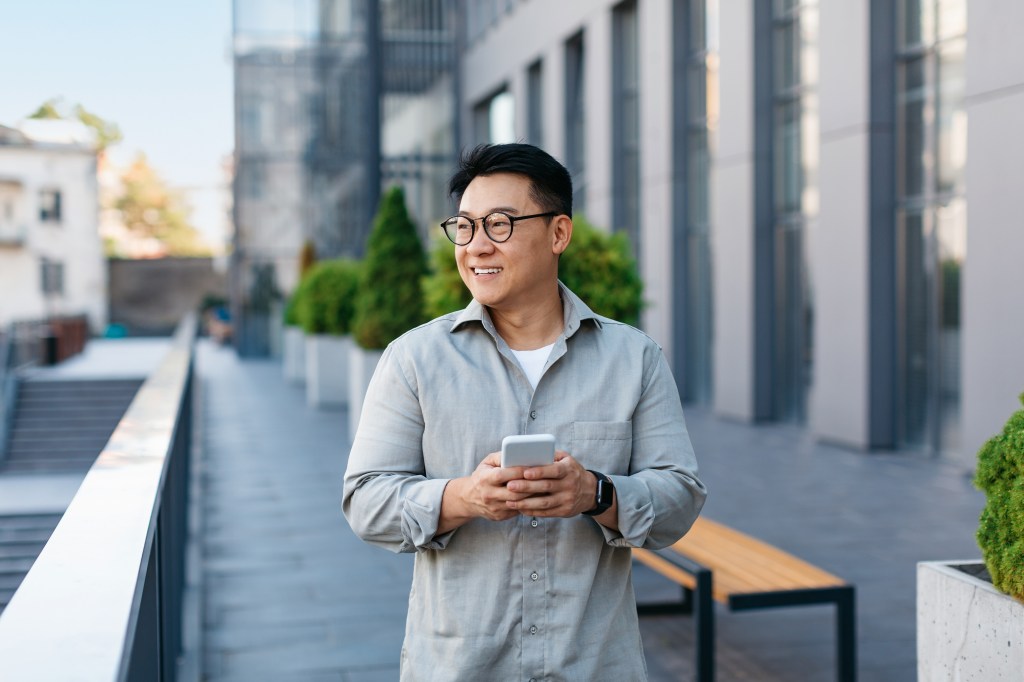 Middle aged male entrepreneur checking emails on smartphone while having break outdoors, walking by business center.