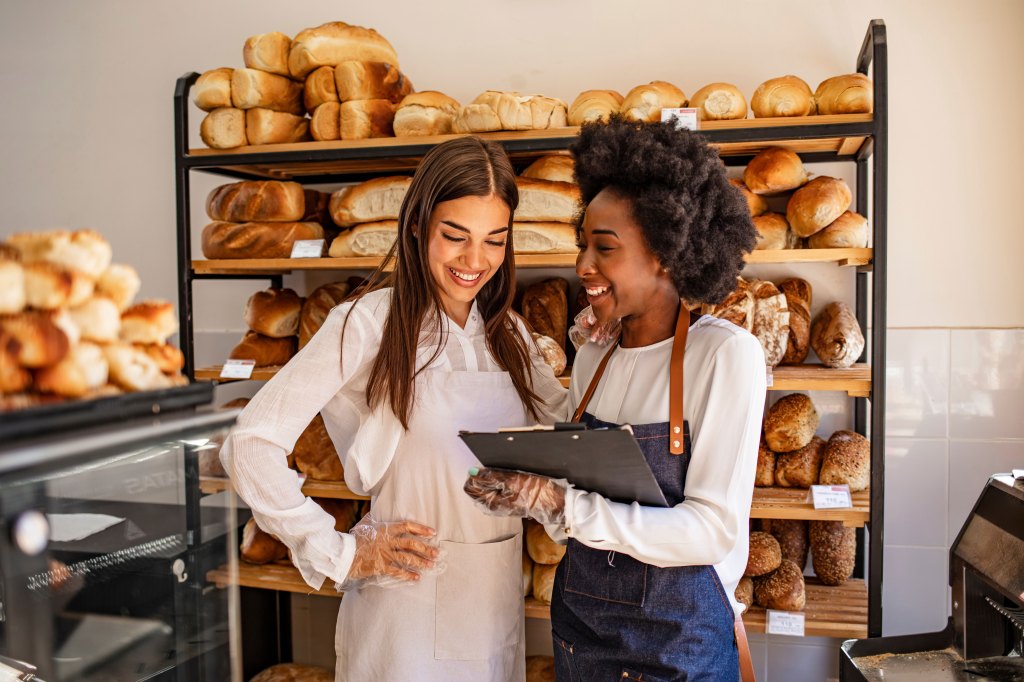 A baker shows her colleague something on her clipboard while they stand in their bakery shop.