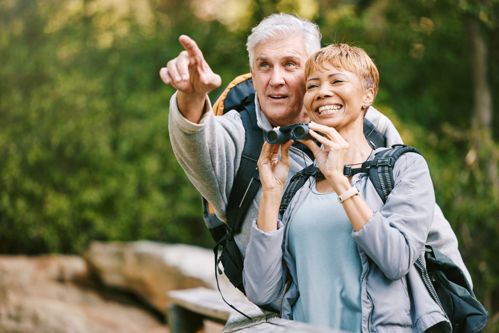 Man and wife pause on a nature walk to bird watch. Female holds a pair of binoculars while the male points to the right.