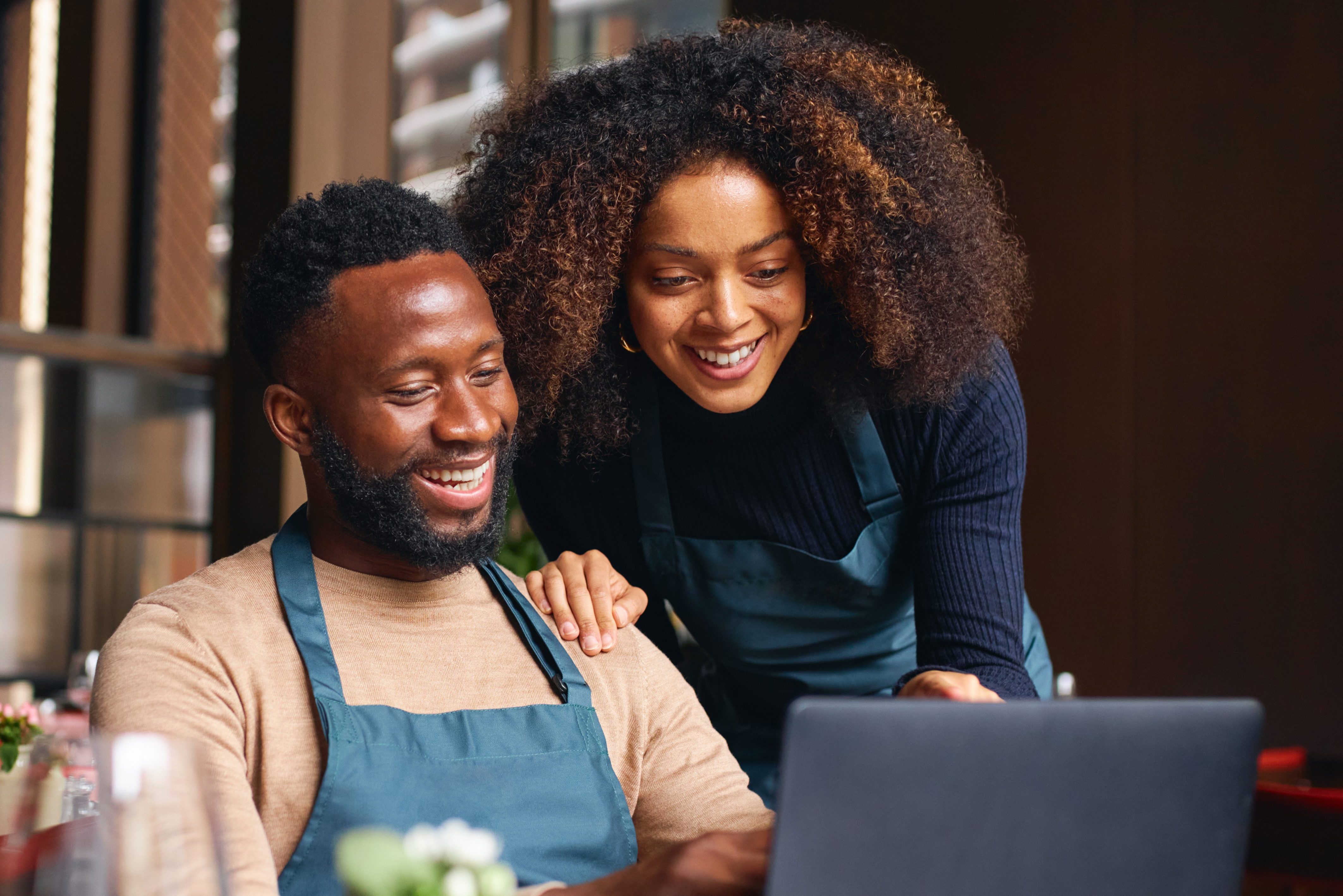 A small business owner sits in a chair at their restaurant while a co-worker shows them business plans on a laptop.