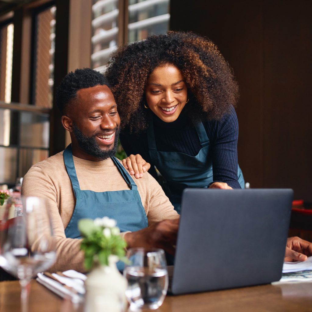 A small business owner sits in a chair at their restaurant while a co-worker shows them business plans on a laptop.