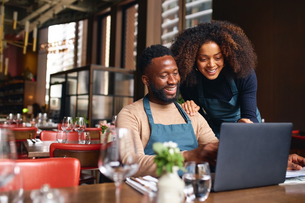 A small business owner sits in a chair at their restaurant while a co-worker shows them business plans on a laptop.