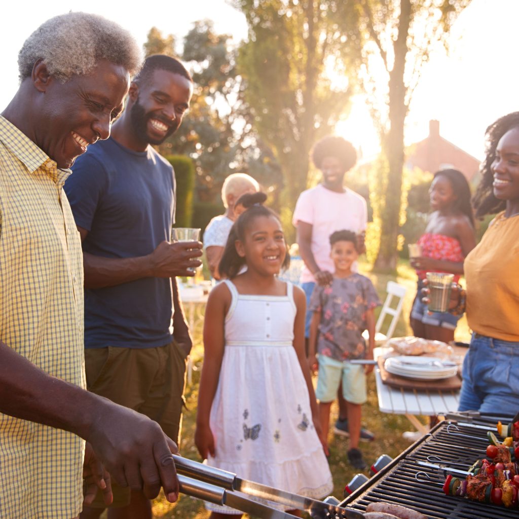 Multi-generational family enjoys an outdoor barbeque. The family stands around the grandfather while he grills.