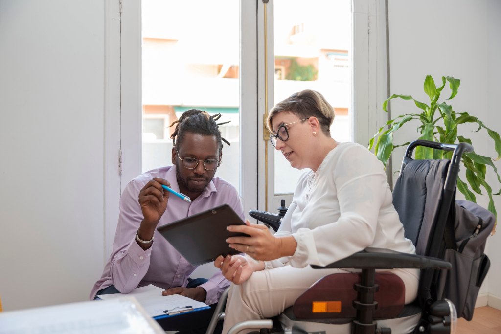 A businesswoman in a wheelchair looks at a digital tablet with her business partner in front of an office window.