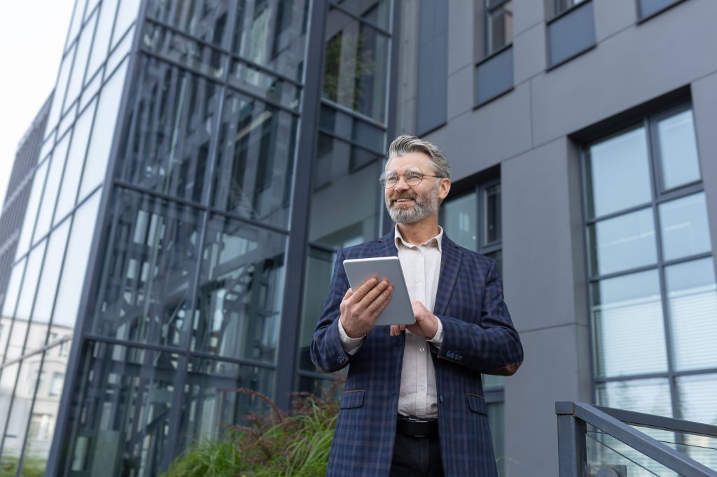 Business man stands outside of commercial building holding a tablet.