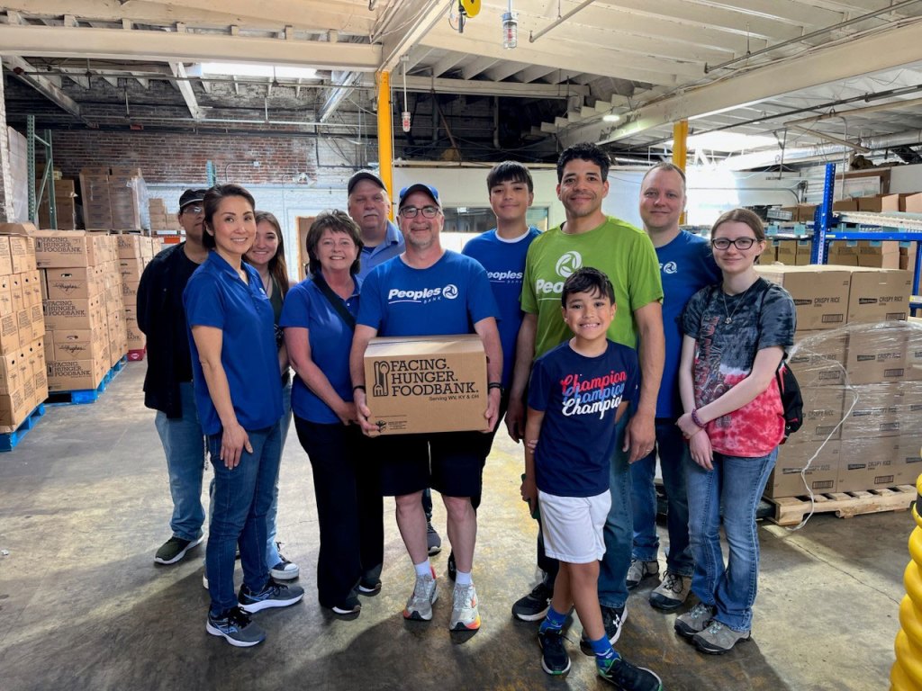 Seven Peoples Bank associates and their children stand in the Facing Hunger Foodbank facility holding food pantry boxes while smiling.
