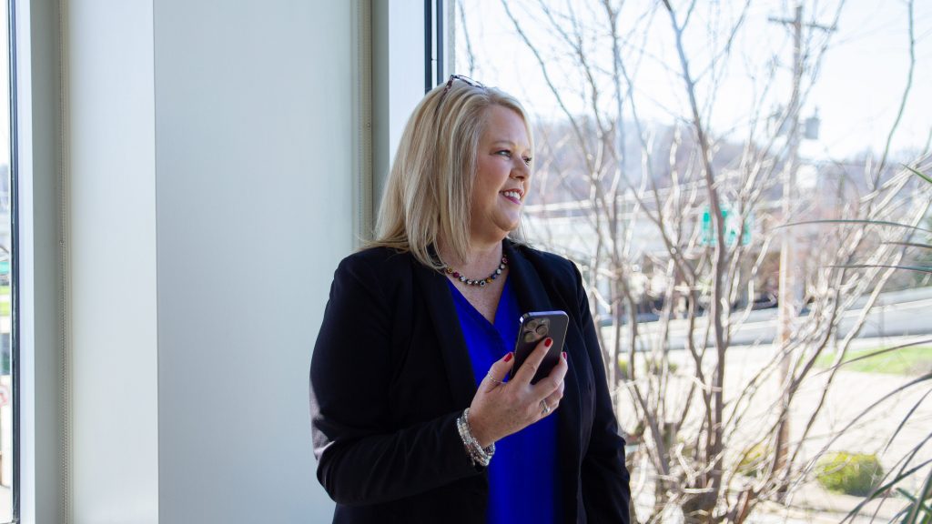 A woman holds a cellphone while looking out bay window onto the city street.