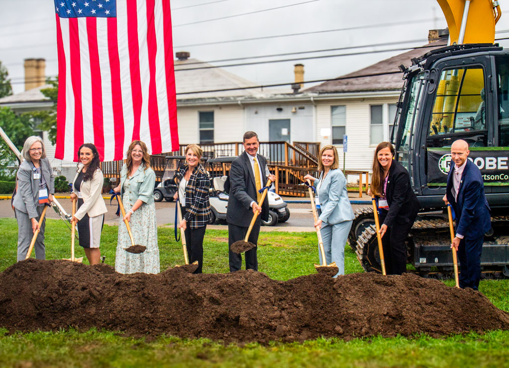 Five Peoples Bank associates join three Marietta Memorial Health Systems employees in the groundbreaking event for the Women and Children's care center in Sistersville, West Virginia. Each person holds a shovel while standing in front of construction gear and an American flag.