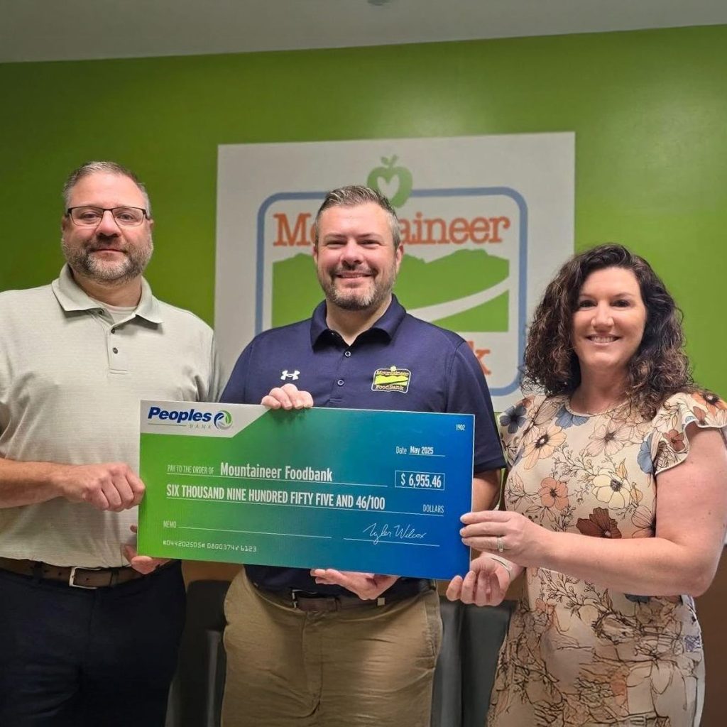 A Peoples Bank associate (left) donates a check from the Peoples Bank Foundation to two Mountaineer Foodbank associates (right). They stand in front of a Mountaineer Foodbank wall logo in a room.