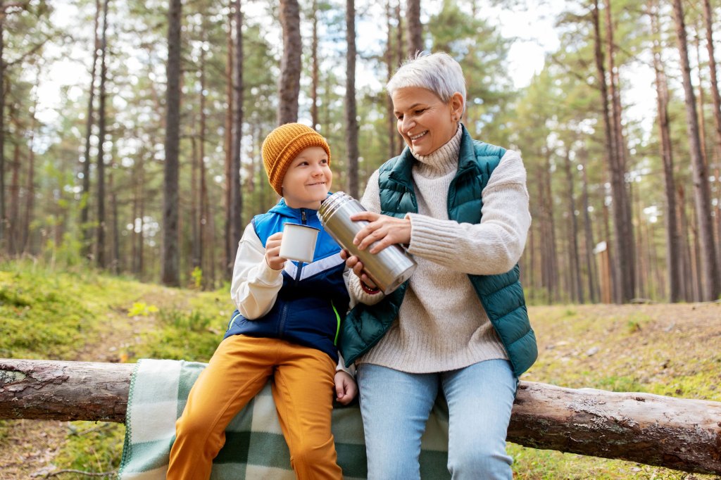 A grandmother (right) and grandson (left) have a picnic on a tree log, drinking hot chocolate.