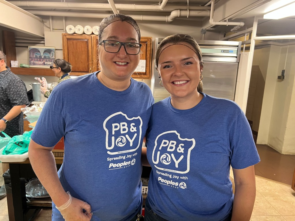 Two Peoples Bank associates stand in a soup kitchen wearing PB & Joy shirts.