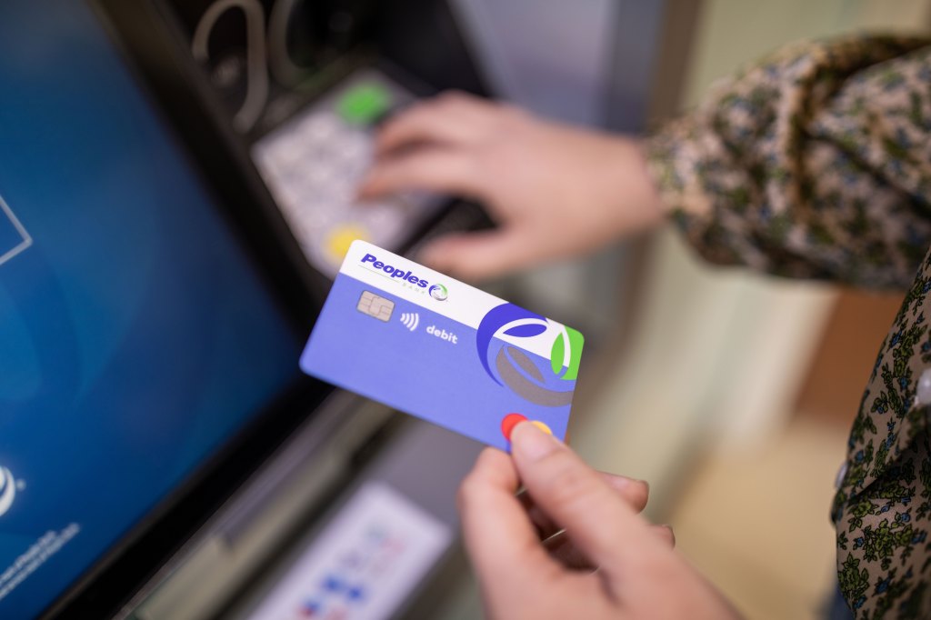 A person holds a Peoples Bank debit card in their left hand while they use a Peoples Bank ATM.