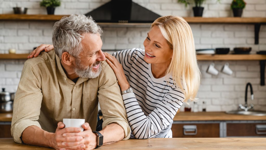 Cheerful senior couple stand in their kitchen while sharing a cup of coffee.
