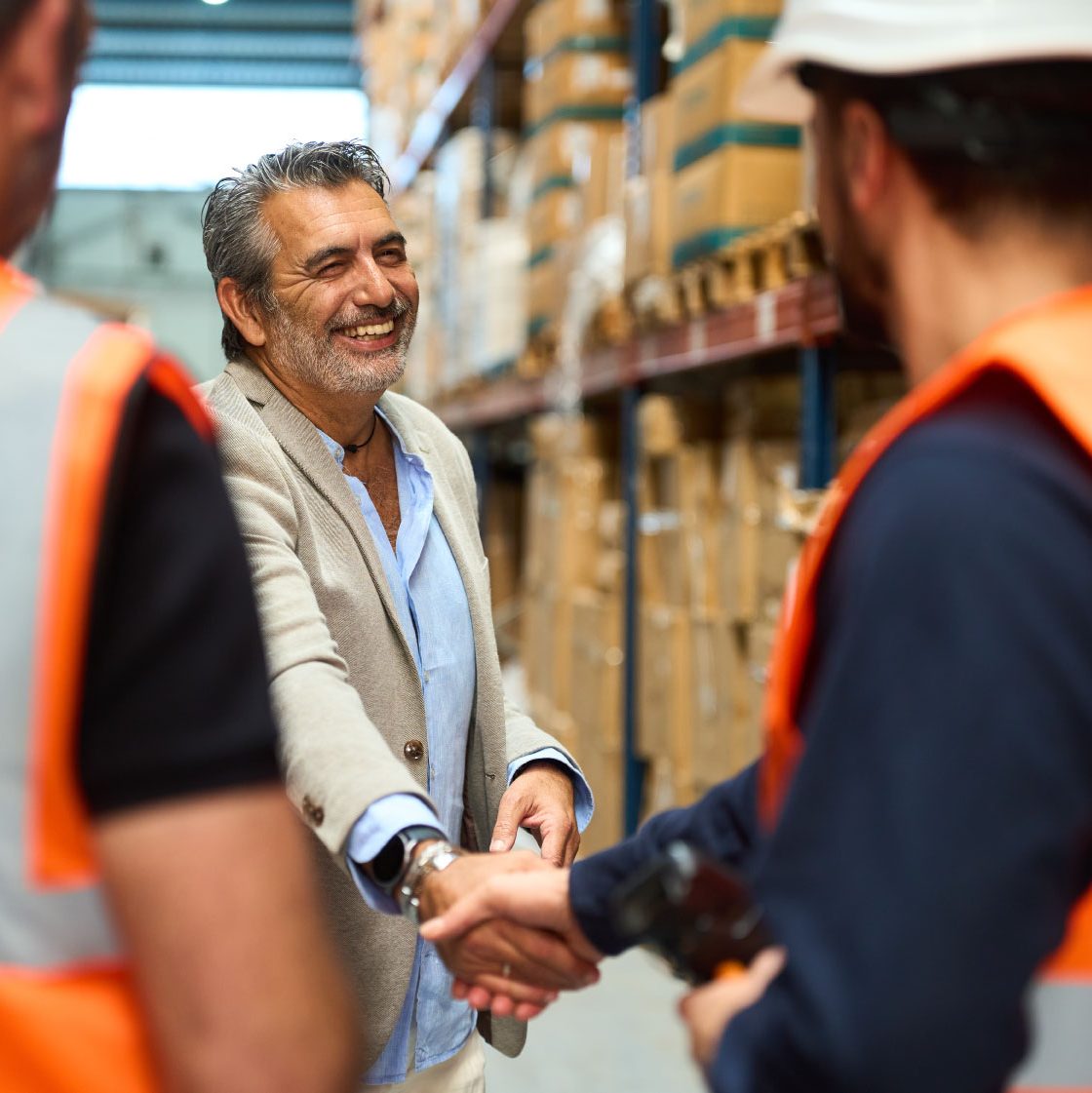 A business manager shakes hands with a warehouse employee dressed in a safety vest and hardhat while standing in a boxing facility.