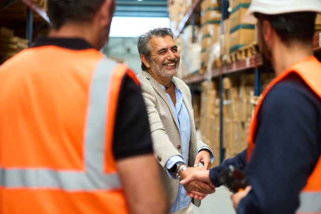 A business manager shakes hands with a warehouse employee dressed in a safety vest and hardhat while standing in a boxing facility.