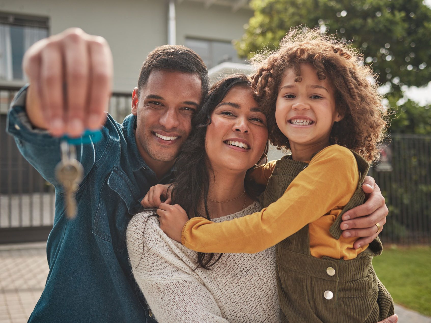 A happy family portrait of a father, mother and child in front of their home. The father holds up the house keys.