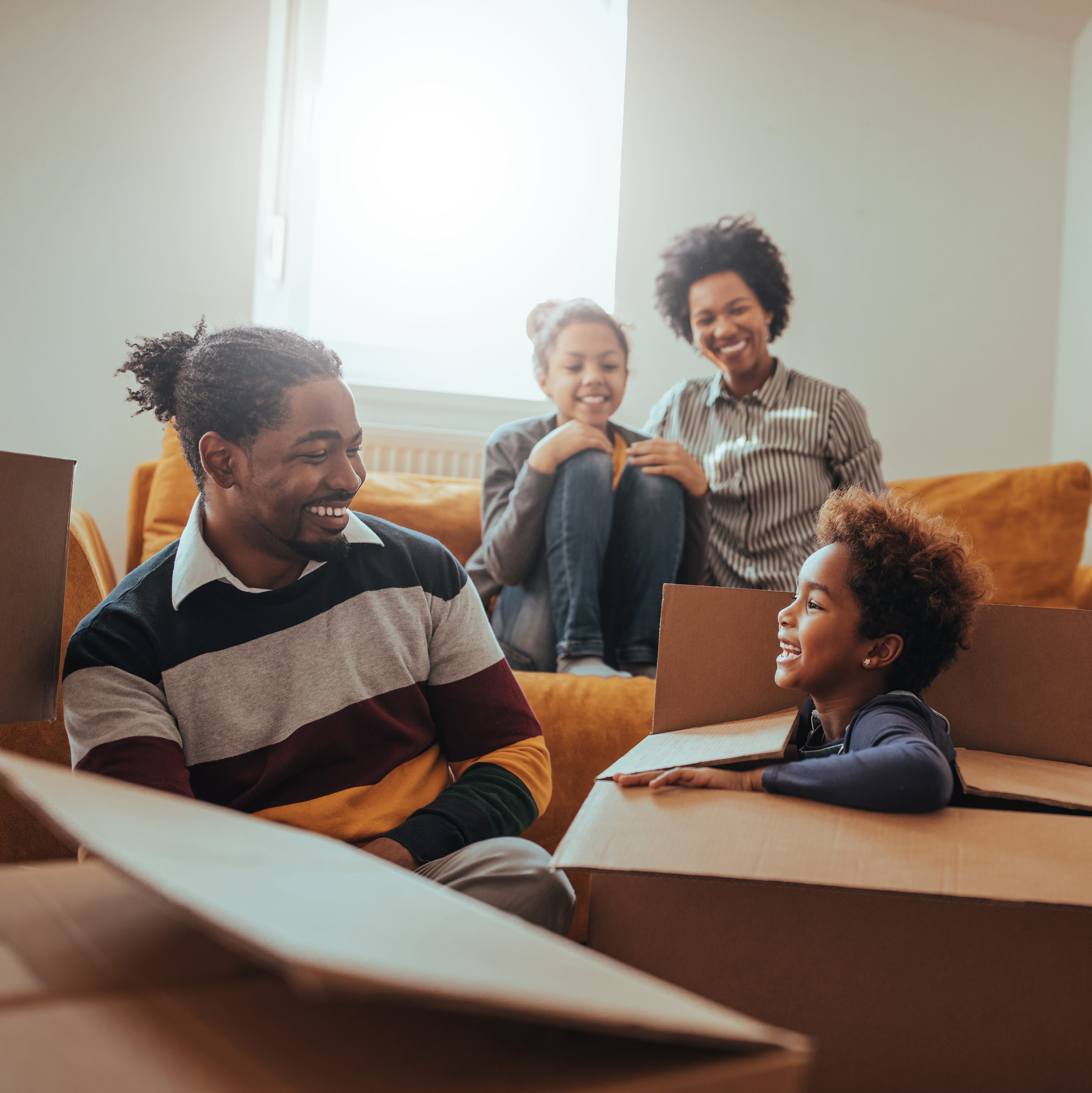 A wife and daughter sit on the couch, watching a husband and son unpack boxes in a new home.