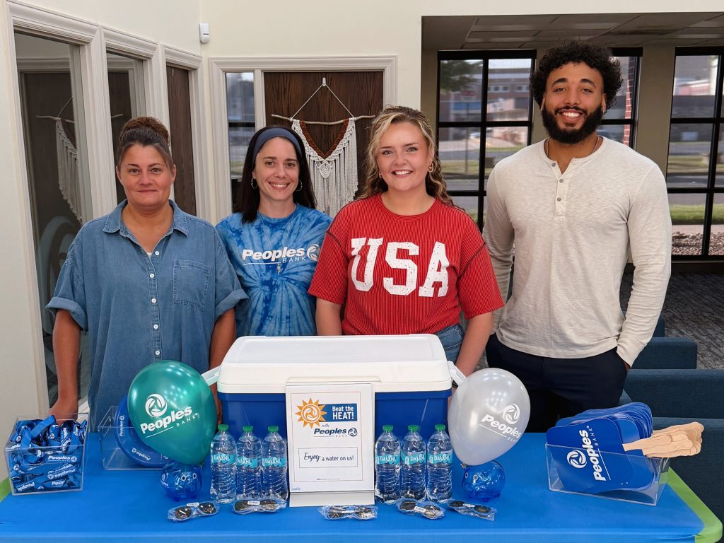Four Peoples Bank associates stand with a table filled with water and fans, welcoming clients to cool down during the summer heatwave.