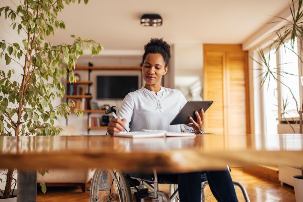 A woman in a wheelchair sits at her dining room table while looking at an ipad and writing in a notebook.