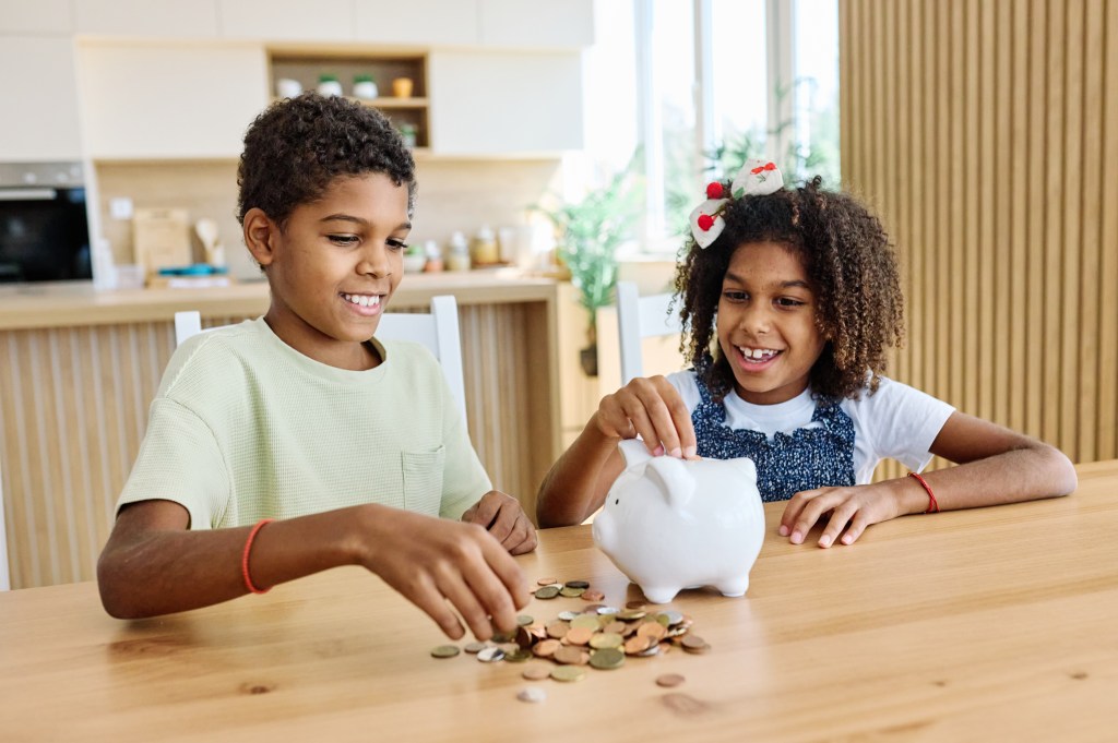 A young boy and his sister put coin money into a piggybank while sitting at the kitchen table.