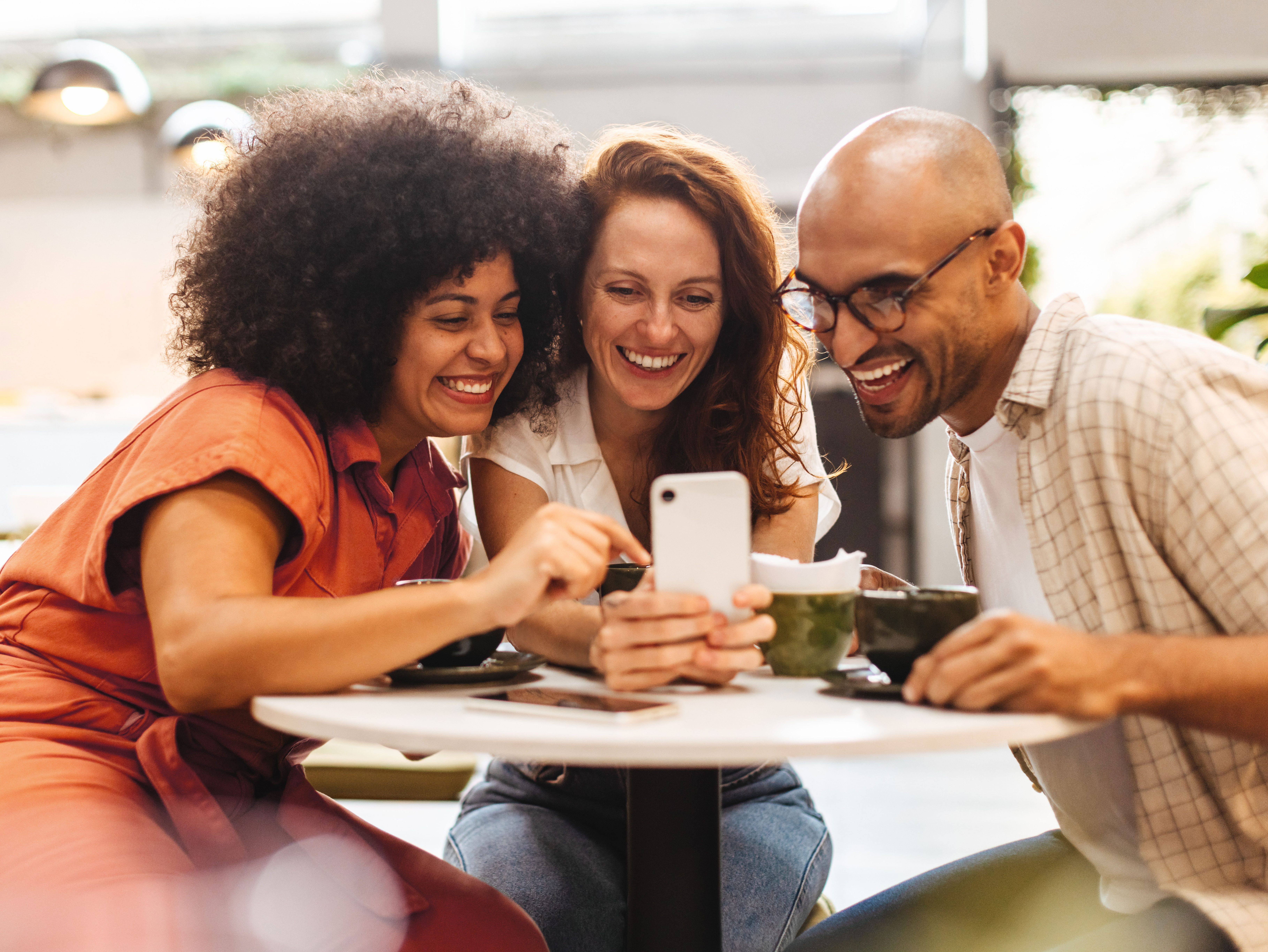 A group of friends sits at a coffee shop while looking at pictures on a phone.